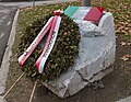 Ein heller großer Stein am Straßenrand mit einer darauf montierten Gedenktafel in Form einer Tricolore und erklärendem Text im mittleren weißen Feld; an der schrägen Front des Steins lehnt ein Gedenkkranz mit Stützen und rot-weißem Schmuckband.